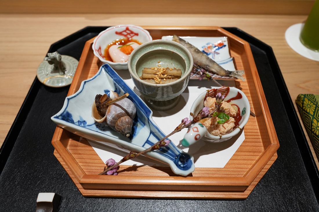 桜の枝をあしらった膳に、磯つぶ貝の旨煮、鯛の子の生姜煮、新牛蒡のたたきを盛った八寸