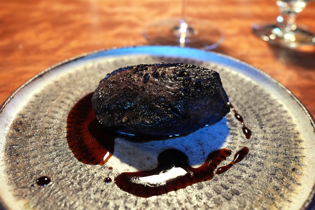 陶器の皿にのせられた、表面を炭火で焼いた黒いめぐみ鴨のかたまり