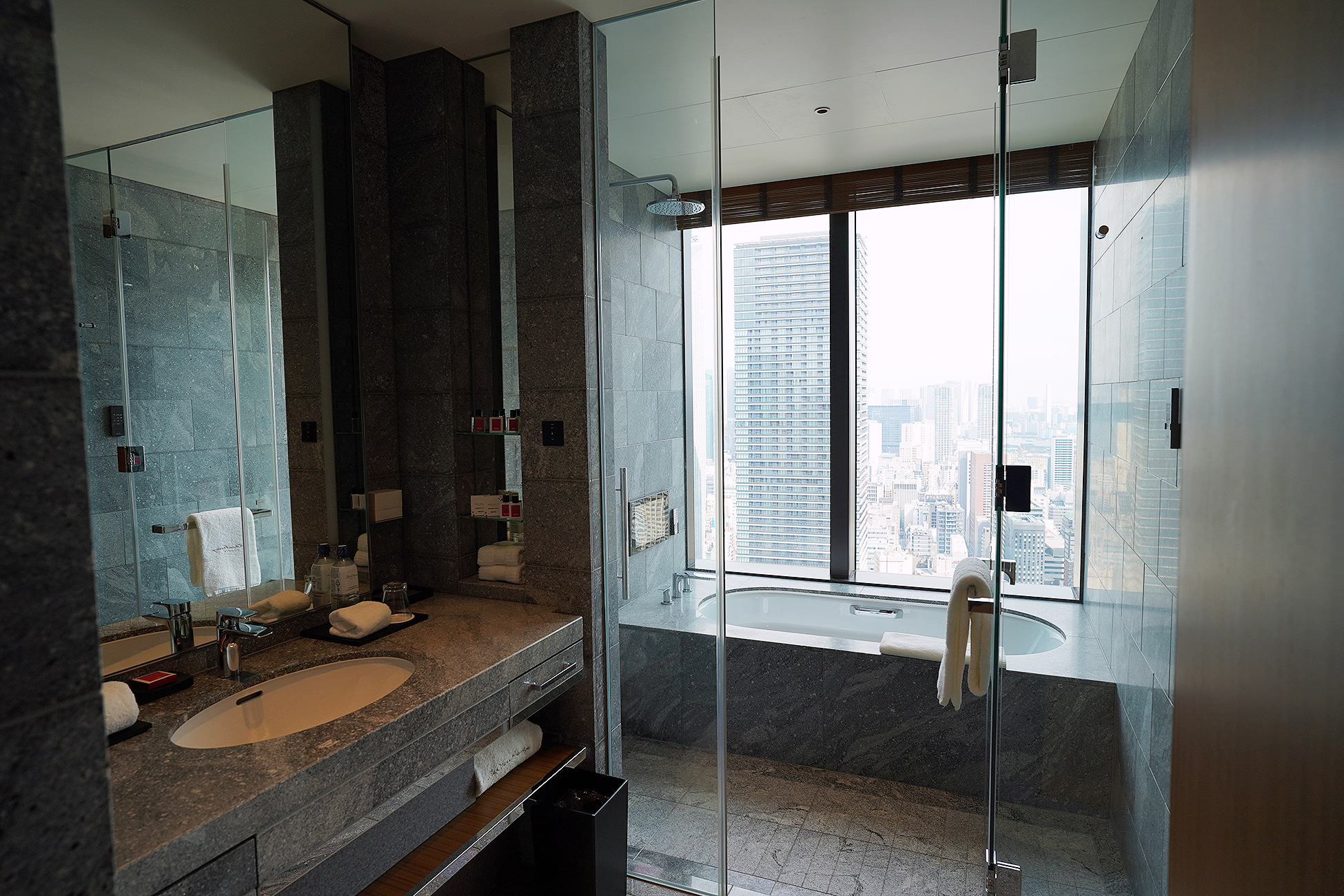 The Okura Tokyoプレステージルームの水まわり全体。黒い石張りの空間に洗面台とバスルームが並び、窓の外にビル群が見える