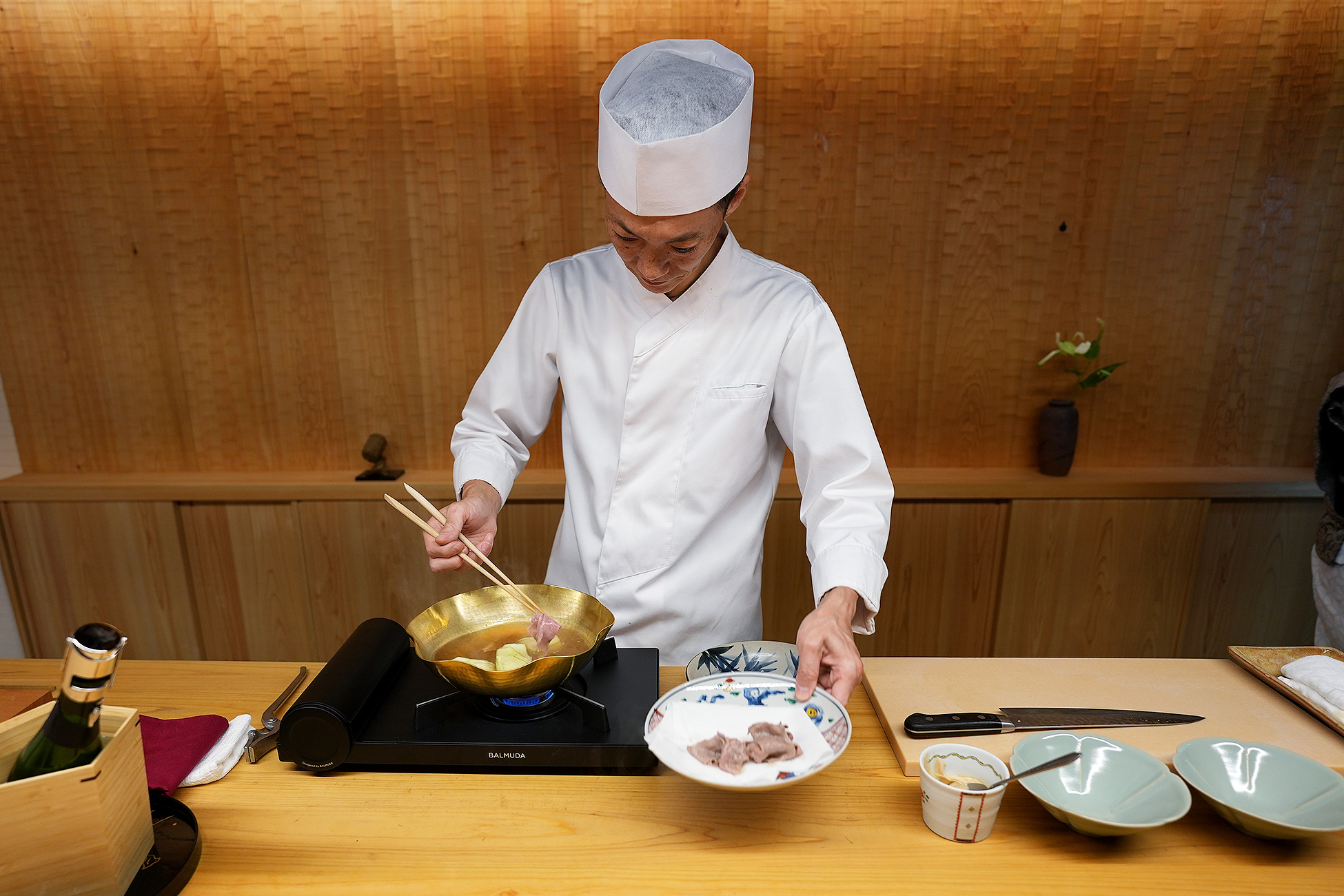 白い調理服と帽子の料理人が、カウンターの上の金色の鍋で肉をしゃぶしゃぶしているところ