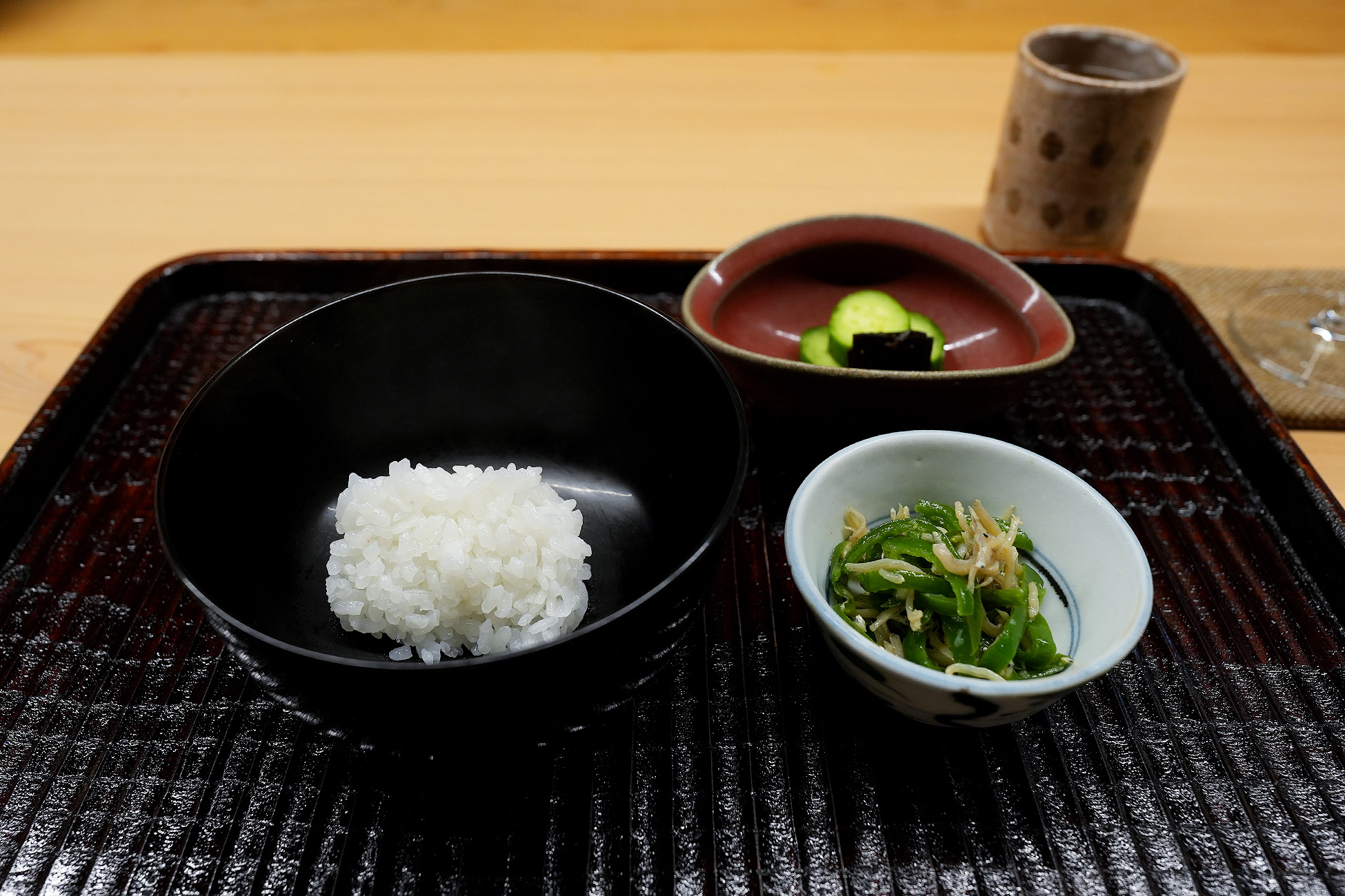 黒い椀によそった白米と、ピーマンとシラスの小鉢、胡瓜と茄子の香の物