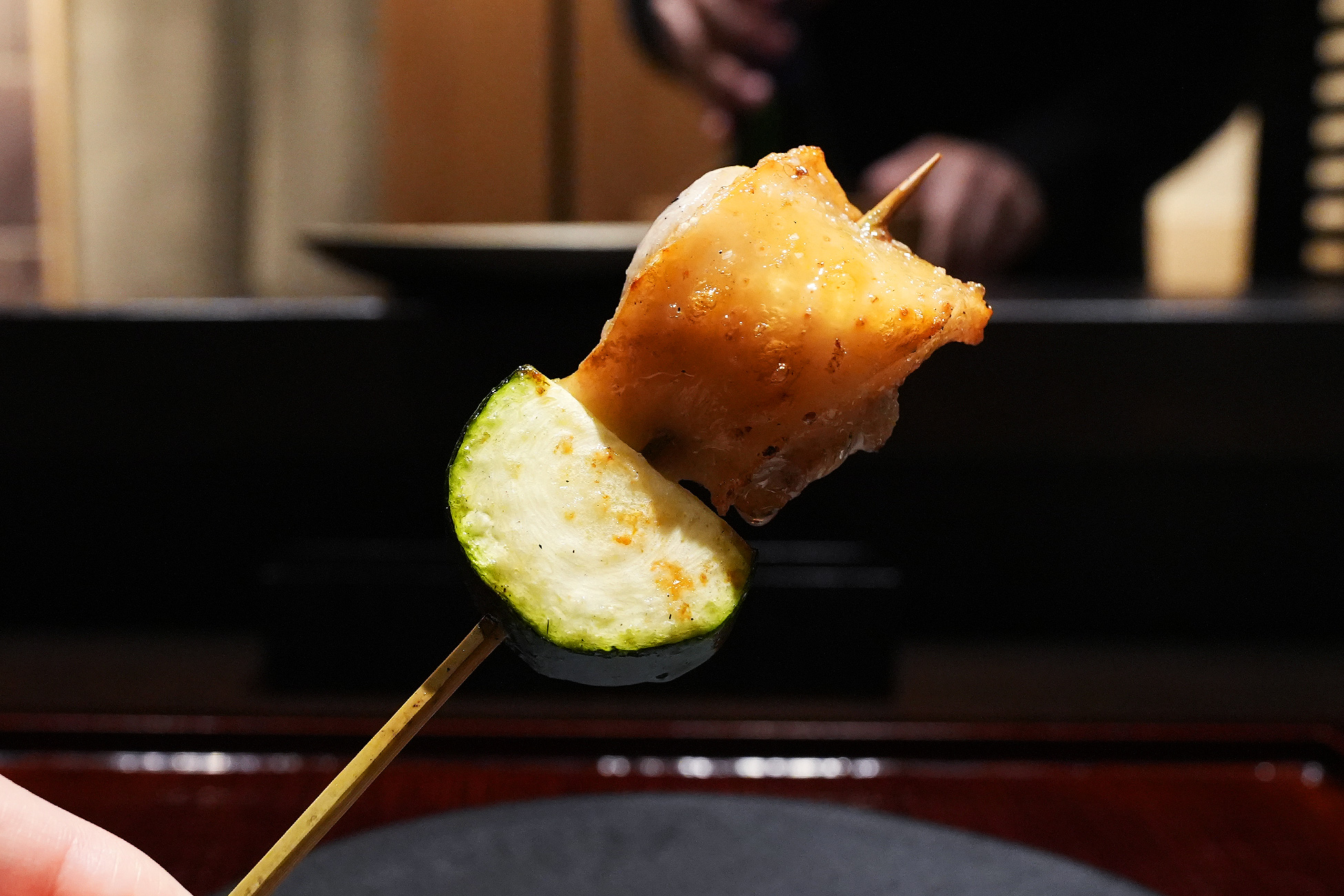 手に持った串。炭火で焼いたもも肉の下に、輪切りのズッキーニが刺さっている