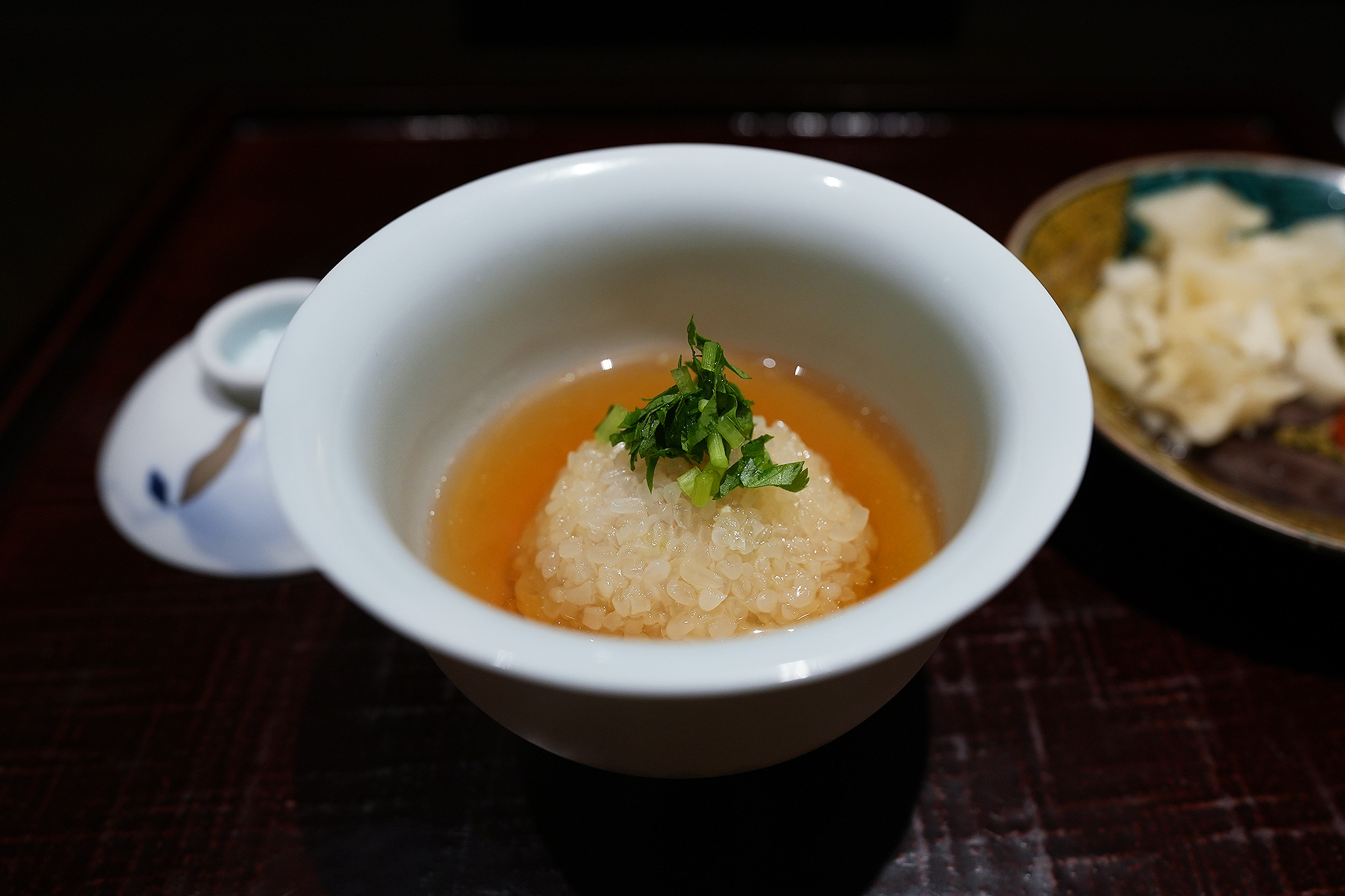 白い鉢に張った琥珀色の出汁。中央に道明寺粉で包んだつくねがあり、上に刻んだ青菜がのっている