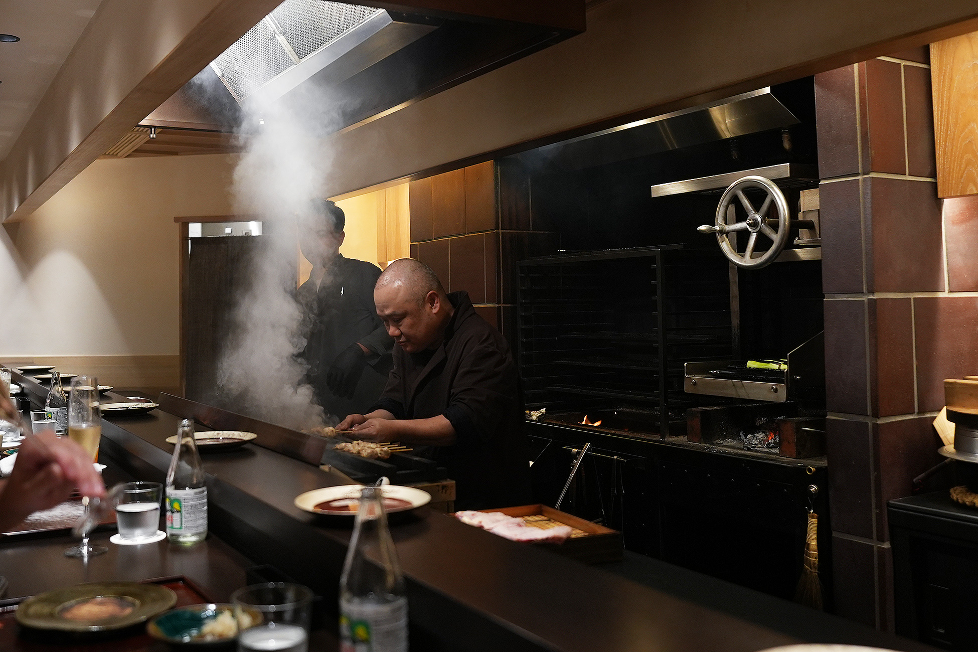 焼き場から白い煙が立ち上がり、カウンター越しに店主が串を焼いている。手前にはグラスやボトルが並ぶ