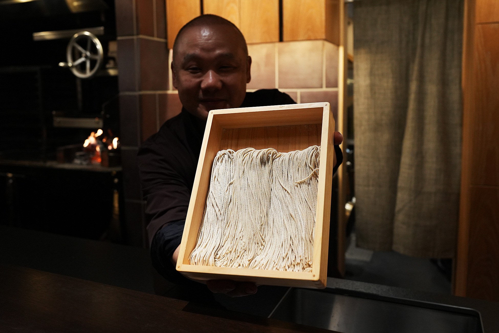 店主が木箱に入った手打ち蕎麦をカウンター越しに見せている。細く切り揃えられた蕎麦が箱いっぱいに並ぶ
