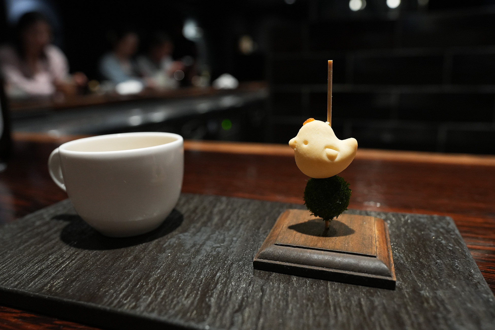 ひよこの形をした皮を串に刺し、苔玉の上に立てた鶏のリエット。木の台にのせられ、左には白いカップが置かれている
